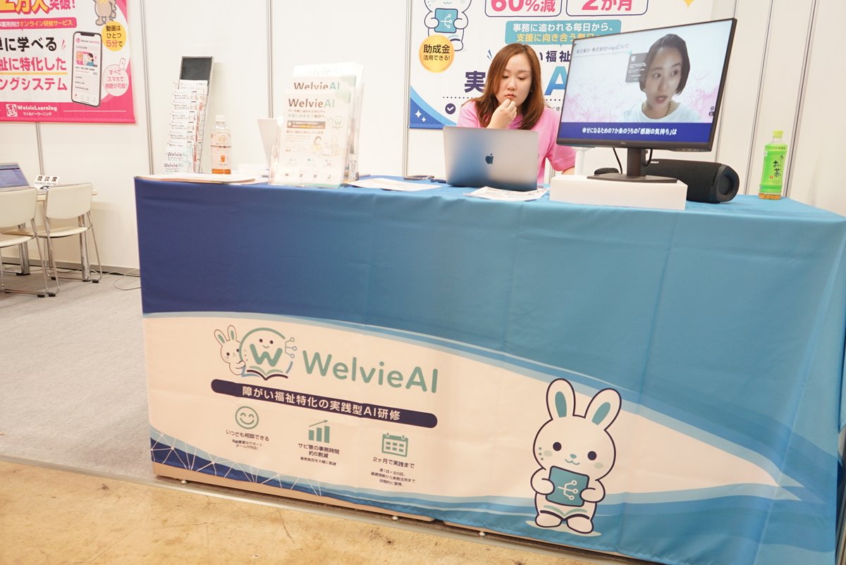 WelvieAIの展示台でパソコンを操作するスタッフとデモ画面